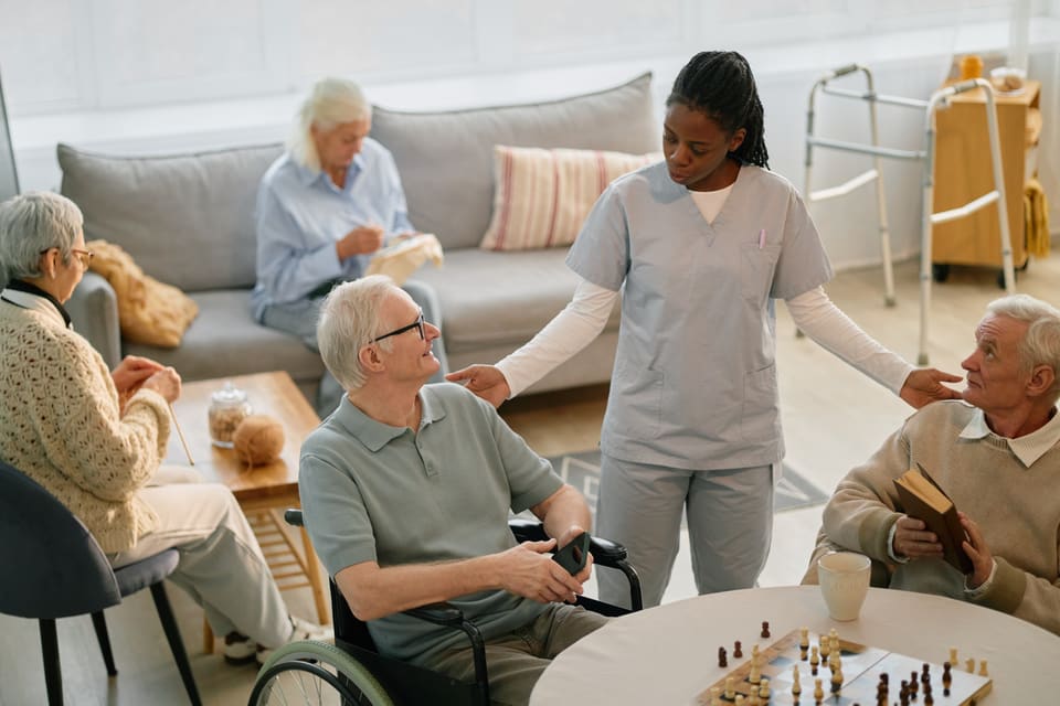 A carer checking in with residents in an aged-care lounge while they play chess and knit