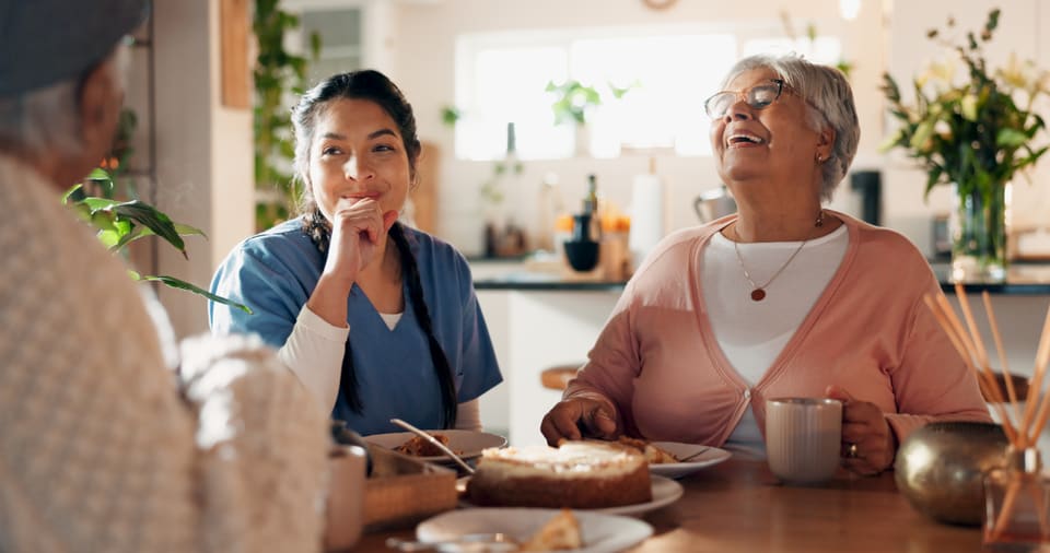 A care worker in scrubs sharing a laugh with two older women over tea and cake at home