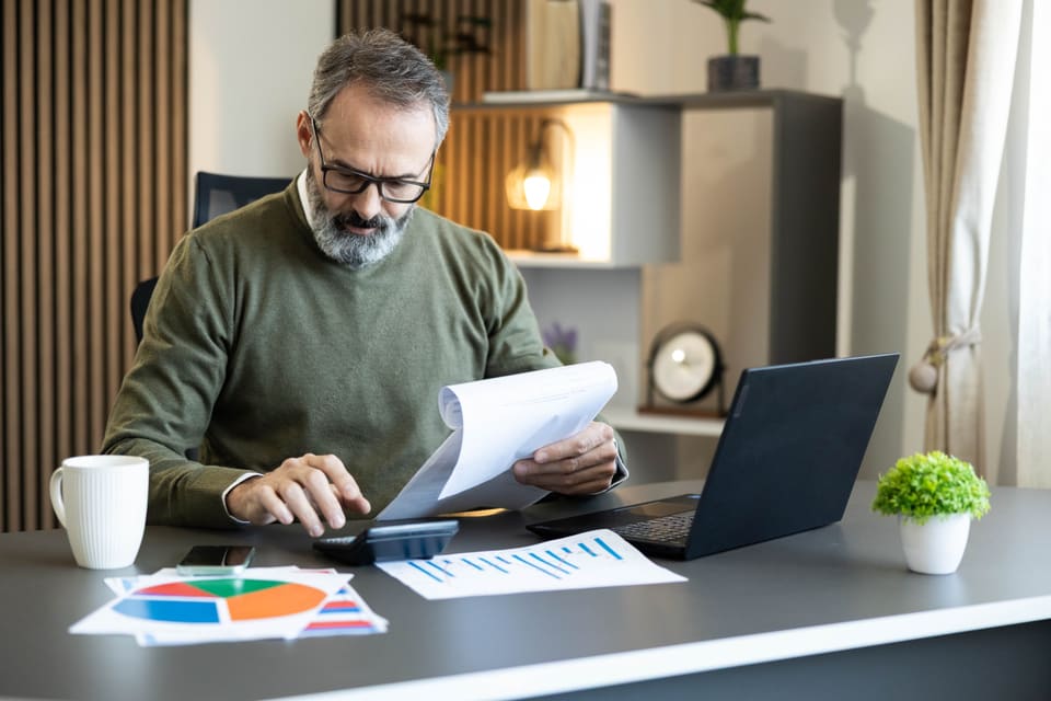 A man at a desk working through figures with a calculator, a printed sheet and his laptop
