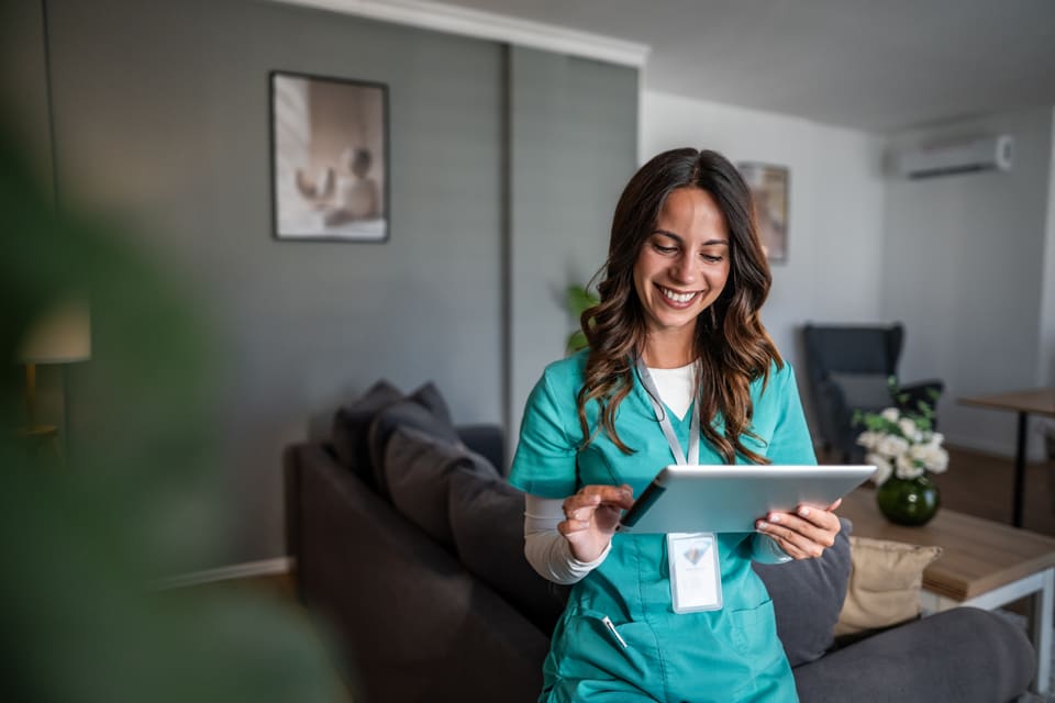 A care worker in teal scrubs smiling as she uses a tablet in a client's living room