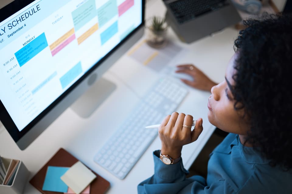 A woman at a desktop computer, hand at her chin, reading a weekly schedule on the screen