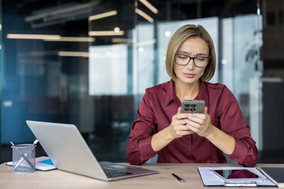A woman at her office desk frowning slightly as she reads something on her phone