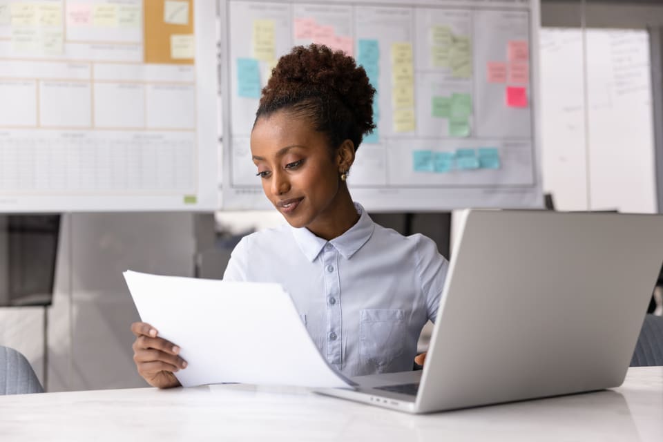 A woman at a laptop reading a printed form, a wall planner behind her