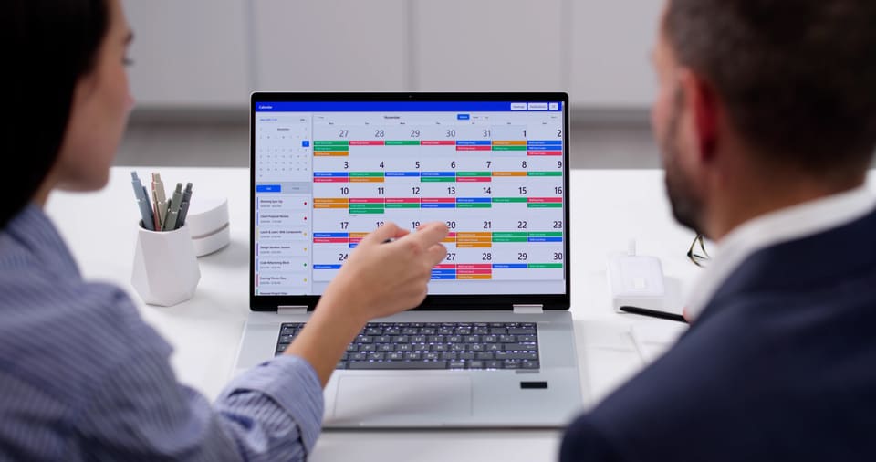 Two colleagues at a laptop, one pointing at a colour-coded monthly calendar on the screen