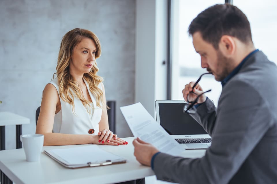 A man reading through a printed page at a desk while the woman across from him waits for his answer