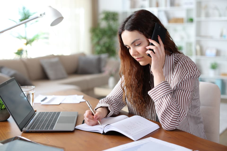 A woman on the phone at a desk at home, writing in a diary