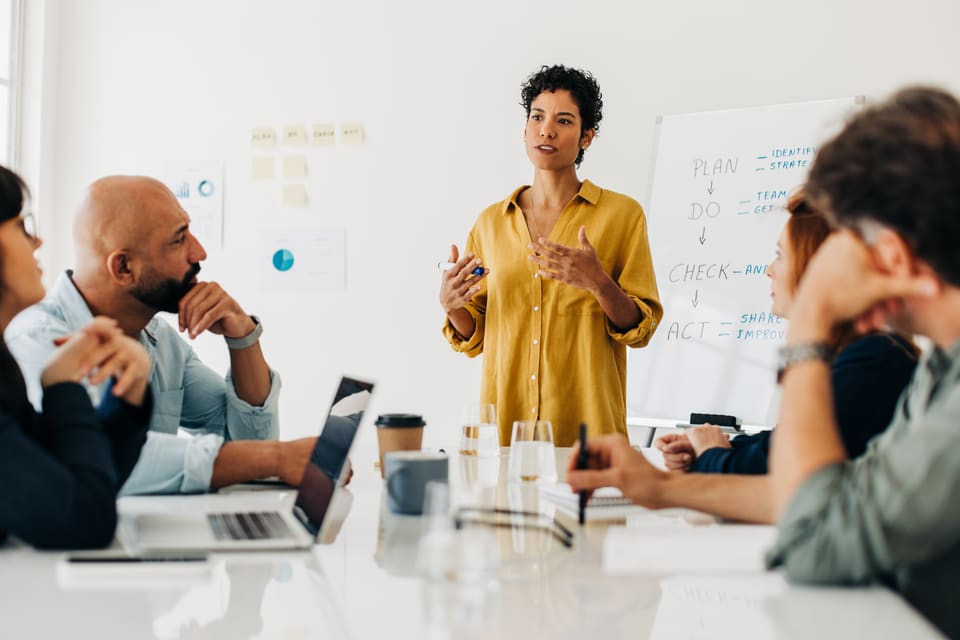 A woman standing at the end of a meeting table, talking her team through a plan on the whiteboard