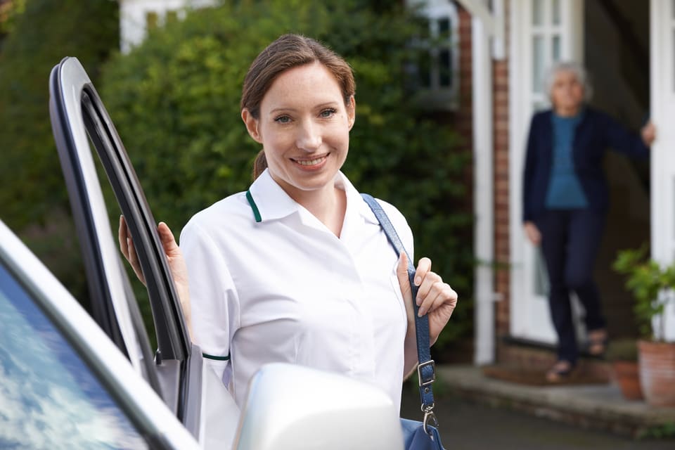 A care worker stepping out of her car at a client's home, with the older woman waiting at the front door