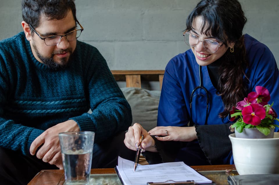A nurse pointing to a line on a form while a man at the kitchen table reads along