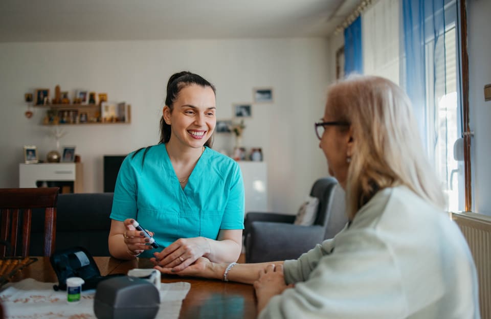 A nurse in teal scrubs checking an older woman's blood sugar at her dining table