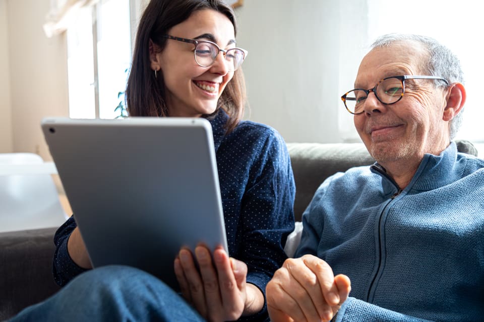 A woman and her older father smiling as they look at a tablet together on the couch