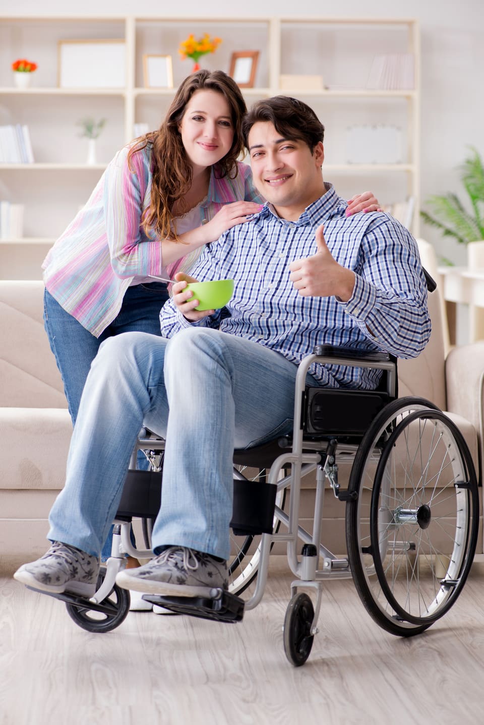 A young woman with her arms around a young man in a wheelchair, both smiling at home