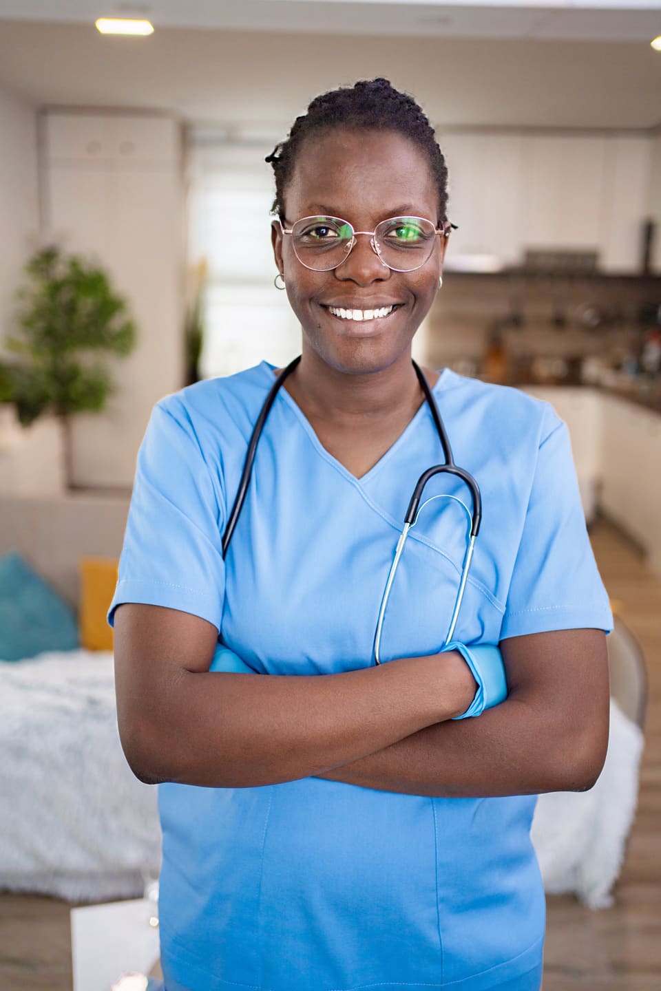 A nurse in blue scrubs with a stethoscope, arms folded, smiling in a patient's home