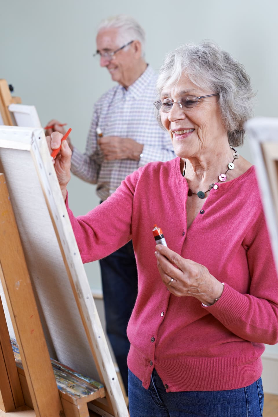 An older woman painting at an easel in an art class, an older man working behind her
