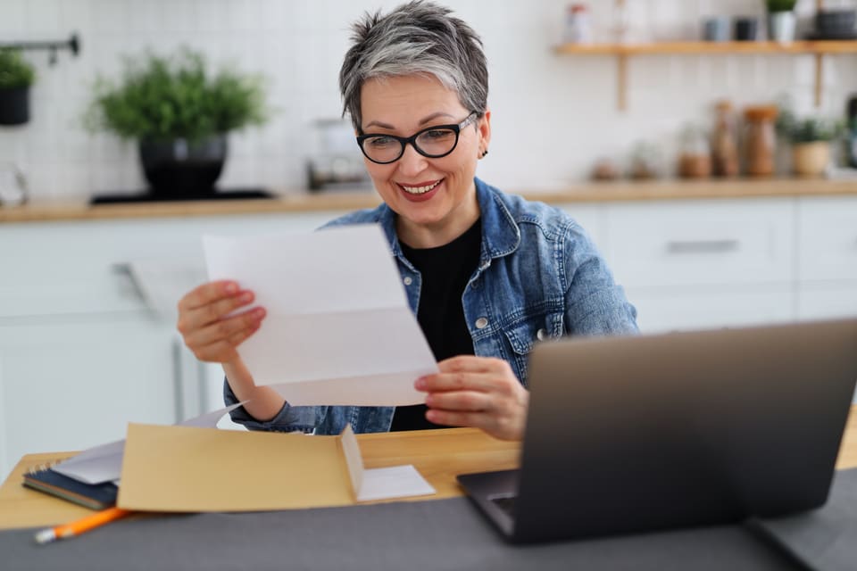 A smiling woman with glasses reading a document at her laptop