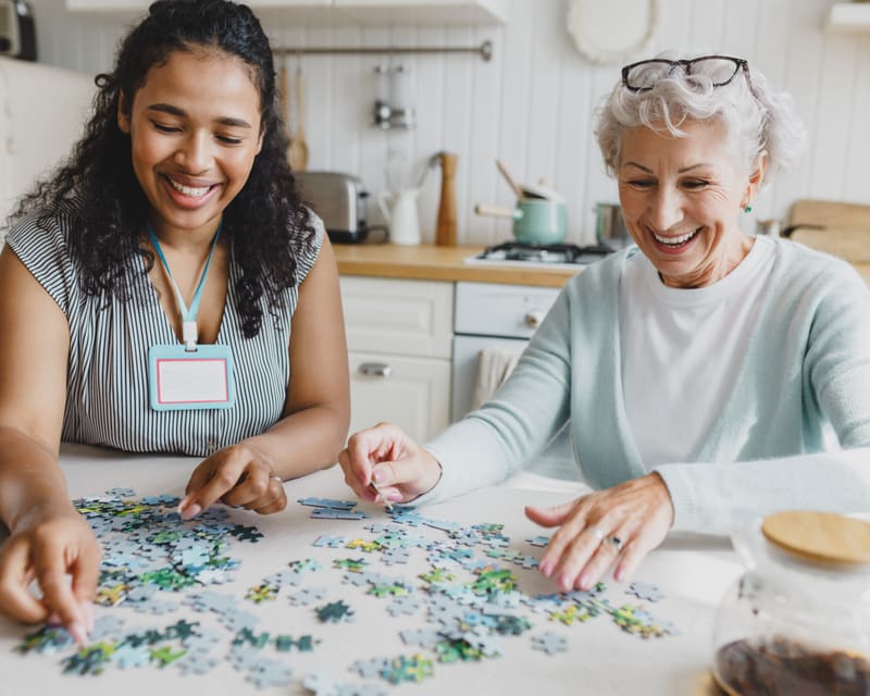 A support worker and an older woman laughing together over a jigsaw puzzle at a kitchen table