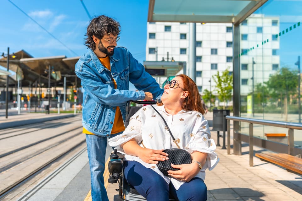 A man pushing a woman in a wheelchair past a tram stop