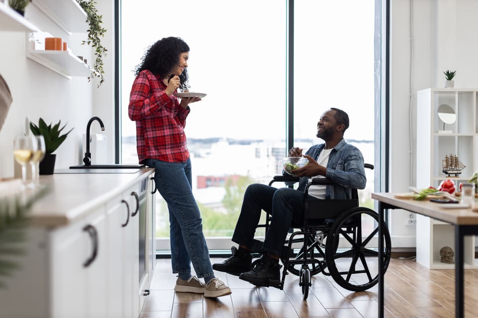 A woman and a man in a wheelchair chatting over coffee in their kitchen