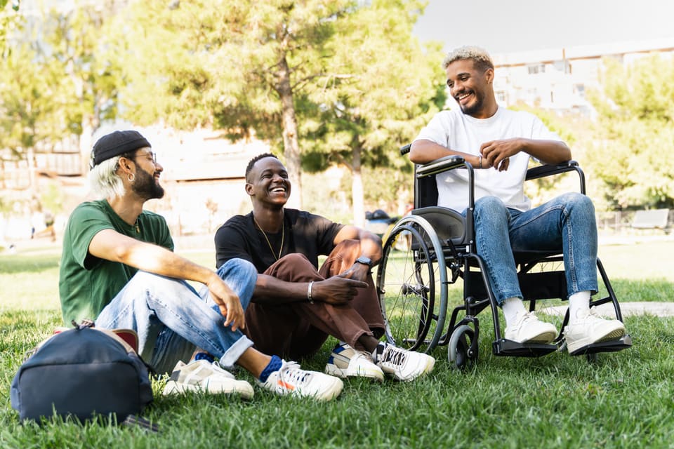 Three friends laughing together on the grass in a park, one of them in a wheelchair