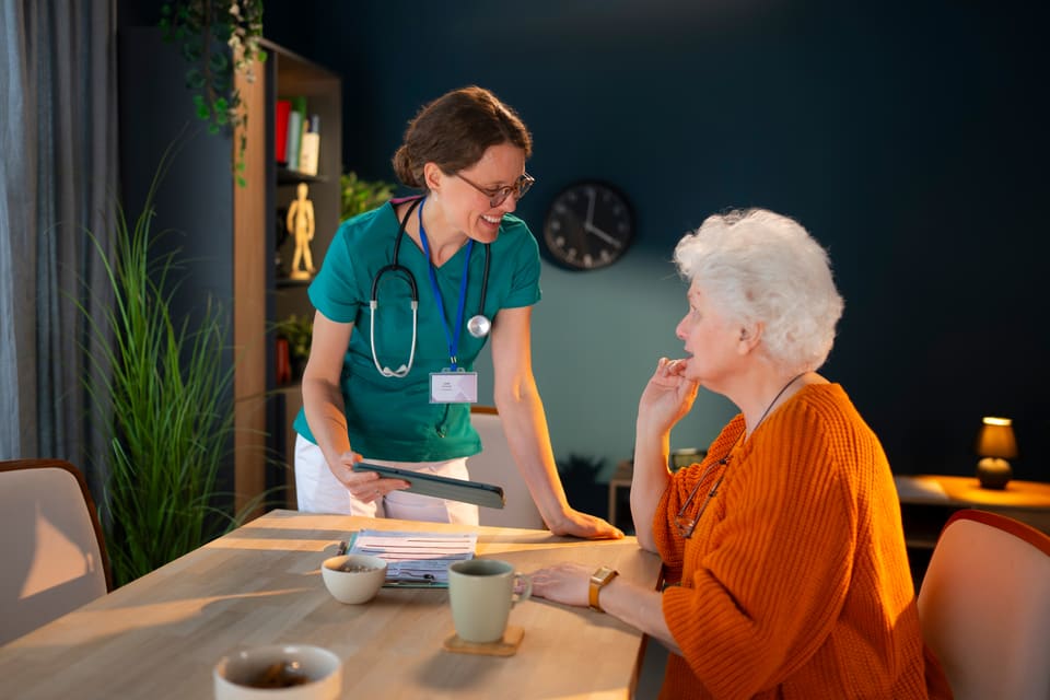 A nurse with a tablet talking with an older woman at her dining table at home