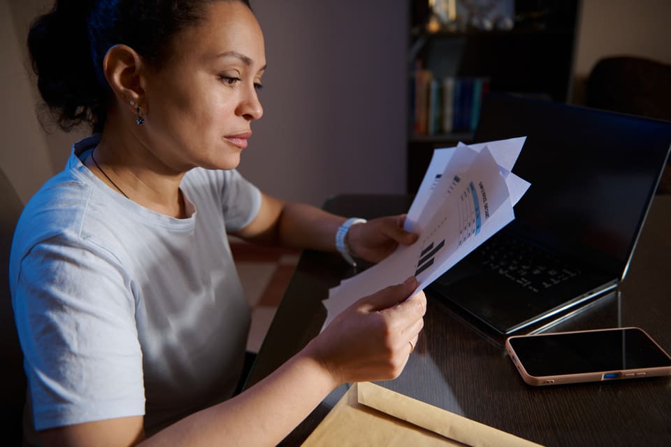 A woman reading through printed reports at her desk in the evening, her laptop beside her