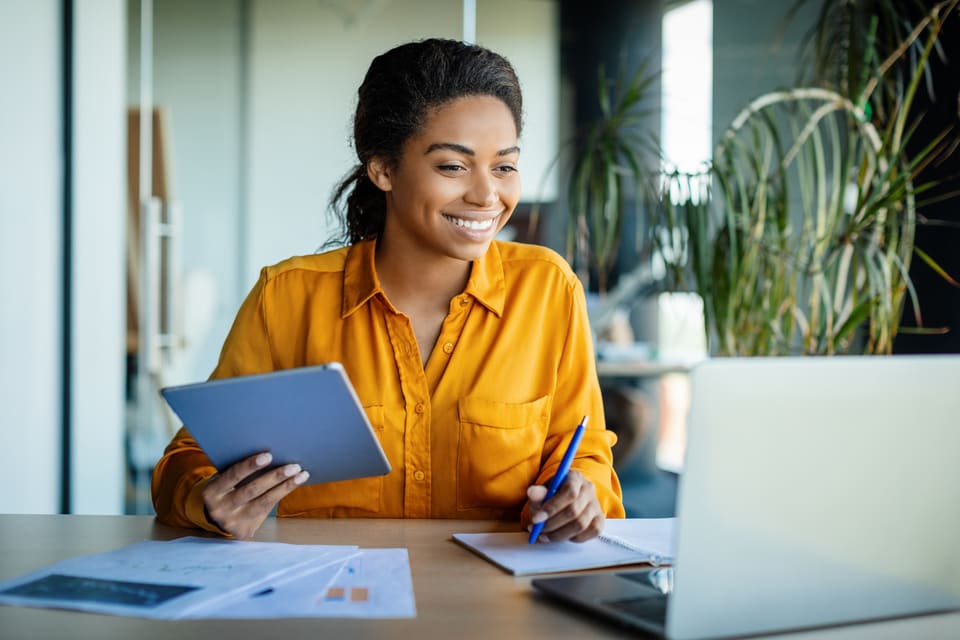 A smiling woman at her desk with a tablet in one hand and a pen in the other, her laptop open beside her