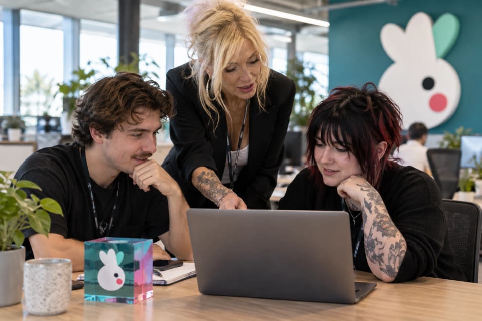 A woman leaning in to point at a laptop between two seated colleagues, a glass Corella block on the desk and the Corella logo on the wall