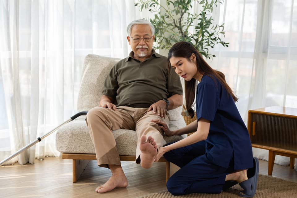 A nurse kneeling to check an older man's foot in his living room